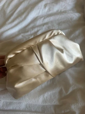 Champagnefärgad satin  clutchbag - Snygg kuvertväska i champagnefärgad satin med mjuk, puffig form och guldfärgad kedja som axelrem. Väskan har ett svart innerfoder och stängs med metallspänne. Perfekt för att lyfta din outfit med en elegant touch.