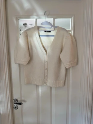 Vit stickad kofta med puffärm - Mysig vit stickad kofta med v-ringning och puffiga korta ärmar. Koftan har knappar framtill och ribbade muddar vid ärmslut och nederkant. Perfekt för lager på lager och enkel att styla till olika outfits.