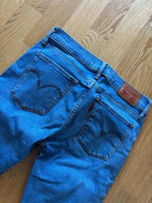 Levi's blå raka jeans - Klassiska blå Levi's jeans i rak modell med hög midja och fem fickor. Jeansen har den ikoniska röda Levi's-lappen på bakfickan och läderpatch bak i midjan. Tillverkade i slitstark denim med snygga kontrastsömmar. Perfekta för en avslappnad och tidlös stil.