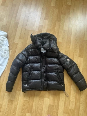 Svart pufferjacka från Moncler - Säljer en svart pufferjacka från Moncler med glansig finish och klassisk quiltad design. Jackan har två dragkedjeförsedda fickor framtill, justerbar nederkant med dragsko och en huva. Moncler-logga på ärmen ger extra stilpoäng.