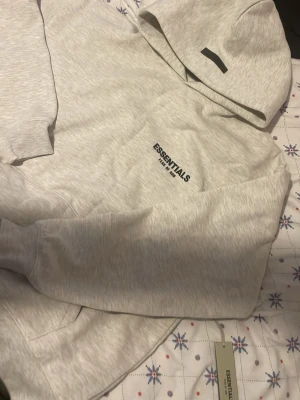  Essentials oatmeal hoodie. - Snygg ljusgrå hoodie från Essentials med svart logga på bröstet. Hoodien har en klassisk huva och känguruficka framtill. Mjuk insida och ribbade muddar vid ärmslut och nederkant. Perfekt för en chill och stilren look.