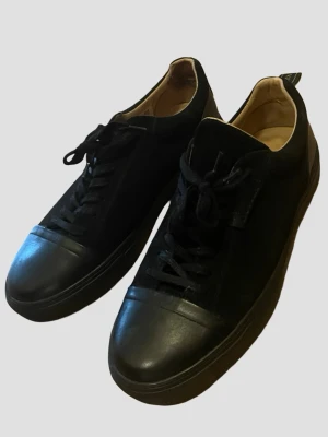Svarta sneakers från Novita - Stilrena svarta sneakers från Novita med ovandel i mocka och tåparti i slätt läder. Skorna har svarta skosnören, låg profil och diskret logga på sidan. Insidan är fodrad i beige läder för extra komfort. Perfekta för en clean och modern look. Nästan helt nya använda 2-3 gånger. 