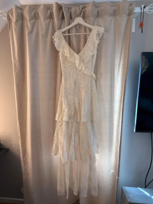 Vit spetsklänning med volanger - Romantisk långklänning i vit spets med v-ringning och volangdetaljer. Klänningen har omlott-design, är ärmlös och har flera lager i kjolen för en flowy känsla. Perfekt för sommarens speciella tillfällen.