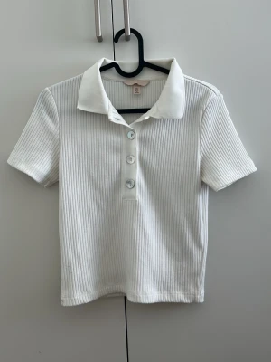Vit ribbad polotopp från H&M - Säljer en vit ribbad polotopp från H&M med korta ärmar och klassisk krage. Toppen har knappar framtill och är tillverkad i mjuk bomull. Perfekt för en clean och stilren look.