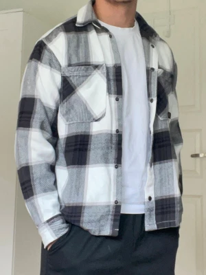 Svartvit rutig overshirt från H&M - Overshirt i svartvitt rutigt mönster från H&M. Skjortjackan har två bröstfickor med lock och knapp, klassisk krage och knäppning framtill. Tillverkad i ett mjukt, tjockare tyg som ger en avslappnad och trendig look.