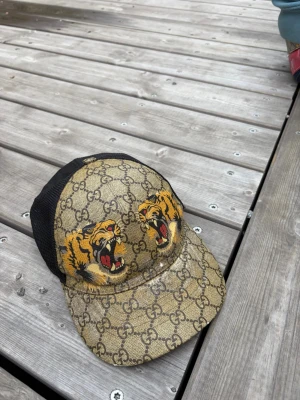 Gucci keps med tigertryck - Säljer en snygg Gucci keps med klassiskt GG-mönster i beige och brunt, prydd med två coola tigerhuvuden framtill. Baksidan är i svart mesh för extra ventilation och kepsen har justerbar rem. Perfekt för dig som vill sticka ut med en unik accessoar.