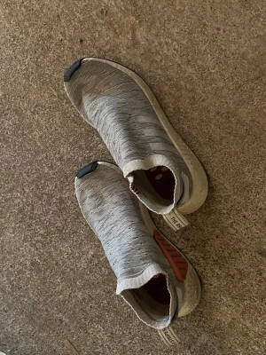 Adidas NMD City Sock grå sneakers - Säljer ett par Adidas NMD City Sock sneakers i ljusgrått med stickad ovandel och vita sulor. Skorna har elastisk strumpliknande passform, svarta detaljer på tån och rosa inslag på sidan. Perfekta för dig som gillar streetwear och bekväma skor.