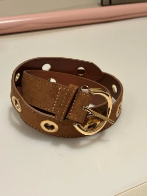 Brunt mockabälte med guldiga hål - Snyggt brunt bälte i mocka med stora guldiga metallhål och matchande guldfärgat spänne. Perfekt accessoar för att lyfta din outfit med en trendig touch. Bältet har bred design och synliga sömmar för extra detalj.