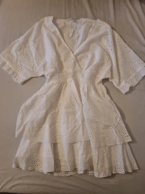 Vit omlottklänning med broderade detaljer - Söt vit kortklänning i bomull med omlottmodell och broderade hålmönster. Klänningen har v-ringning, korta vida ärmar med volangkant och smock i midjan för en snygg passform. Perfekt för sommarens alla tillfällen.