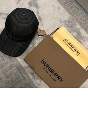 Burberry  keps - Snygg keps från Burberry i klassiskt rutmönster i grått, svart och röda detaljer. Justerbar rem bak och böjd skärm. Tillverkad i bomull och polyester. Perfekt accessoar för dig som vill ha en stilren och exklusiv look. Använd ca 2-3 gånger max pga att den är lite stor för mig. Kommer även med box,  etikett samt en påse till kepsen av burberry.