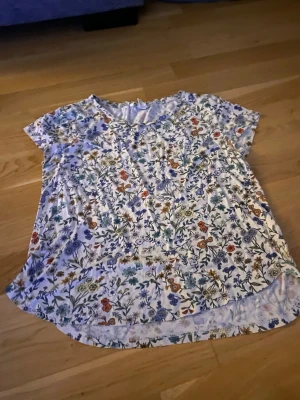 Blommig t-shirt från H&M Basic - Kortärmad t-shirt från H&M Basic med blommigt mönster i flera färger på vit botten. Rund halsringning och avslappnad passform. Perfekt för dig som gillar färgglada och somriga plagg.