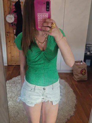 Grön spetsblus med v-ringning - Snygg grön blus i spets med v-ringning och twistad detalj framtill. Kortärmad och figurnära passform som ger en feminin siluett. Perfekt att styla med jeans eller kjol för en trendig look.