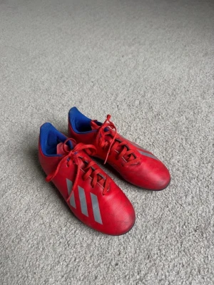 Röda Adidas fotbollsskor - Säljer ett par röda fotbollsskor från Adidas med klassiska tre vita ränder på sidan. Skorna har blå insida och röda skosnören. 