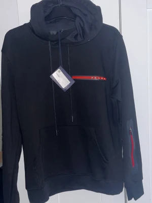 Prada hoodie - Snygg svart hoodie från Prada med röd och vit logga på bröstet. Helt ny. Aldrig använd, endast testad storlek L passar även M. Priset kan diskuteras 