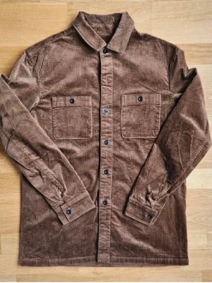 Brun manchester overshirt - Snygg brun overshirt i manchestertyg med två bröstfickor och knappar framtill. Klassisk krage och långa ärmar med knapp vid ärmslut. Perfekt lager på lager-plagg för en avslappnad stil.