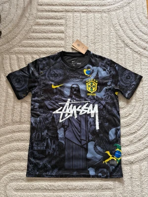 Nike x Stüssy Brasil t-shirt svart - Snygg svart t-shirt från Nike i samarbete med Stüssy och Brasil. Tröjan har coolt grafiskt tryck med Kristusstatyn, Brasil-logga och Stüssy-logga i vitt. Gula detaljer och Nike Swoosh. Perfekt för dig som gillar streetwear och fotboll.