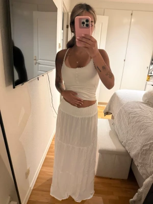 Vit långkjol med volangdetalj - Säljer en vit långkjol med bred smock i midjan och volangdetalj. Kjolen har en luftig och flowy passform, perfekt för varma dagar. Materialet känns lätt och bekvämt, troligen bomull eller polyester. Enkel och stilren design som är lätt att matcha. Storlek xs men passar även S då den är stretchig 