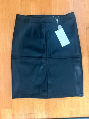 Svart skinnkjol med raka linjer - Snygg svart kjol i skinnimitation med raka sömmar och minimalistisk design. Kjolen har en klassisk, rak passform och slutar ovanför knät. Slits baktill. Perfekt för dig som gillar en stilren look. Liten i storleken, mer en small än medium.