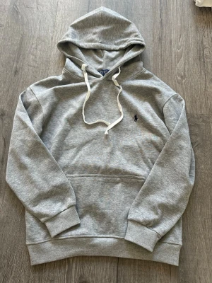 Grå hoodie från Polo Ralph Lauren - Snygg grå hoodie från Polo Ralph Lauren med klassisk huva och vita dragsnören. Tröjan har en broderad marinblå logga på bröstet och en stor magficka framtill. Tillverkad i mjuk bomullsblandning och har ribbade muddar vid ärmar och nederkant.
