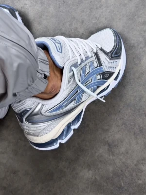 ASICS vita och blå sneakers - Snygga sneakers från ASICS i vitt meshmaterial med blå och silverfärgade detaljer. Skon har en sportig vibe med tydliga logor på sidan och en chunky sula med blå inslag. Perfekt för dig som gillar streetwear och sportig stil.
