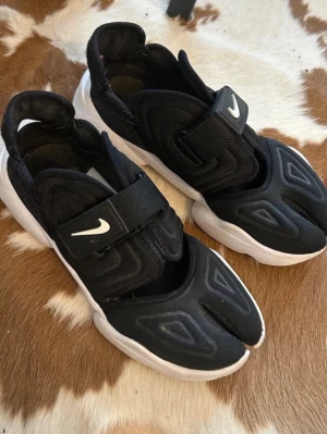 Nike Air Rift svarta sneakers - Unika svarta Nike Air Rift sneakers med delad tå och bred vit sula. Skorna har en sportig look med kardborreband över foten och vit Nike-logga. Ovandelen är i textil och mesh för extra komfort och andningsförmåga. Perfekta för dig som vill sticka ut med din stil. Pris kan diskutera vid snabb affär