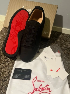 Svarta sneakers från Christian Louboutin - Snygga svarta sneakers från Christian Louboutin med klassisk röd sula och diskret logga. Skorna är låga och har ovandel i mocka med matchande svarta skosnören. Perfekta för dig som vill ha en clean och exklusiv look.