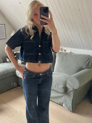 Mörkblå croppad jeansskjorta - Säljer en mörkblå croppad jeansskjorta med korta ärmar och stora bröstfickor. Skjortan har klassisk krage och knäppning framtill med silvriga knappar. Perfekt till ett par höga jeans för en cool denimlook.