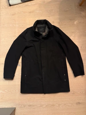 Svart Gore Tex jacka från Peak Performance  - Stilren svart jacka från Peak Performance i GoreTex med hög krage och dold dragkedja. Rocken har raka linjer, två sidofickor och minimalistisk design. Perfekt för dig som gillar en clean och modern look. Finns en huva att ta fram. Storlek XXL