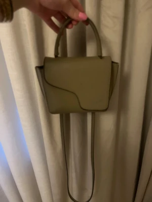 ATP väska  - Snygg olivgrön minihandväska med både handtag och lång axelrem. Väskan har en unik asymmetrisk lockdesign och stilrena detaljer i silver. Perfekt för dig som gillar minimalistisk och trendig stil. Tillverkad i slätt konstläder.