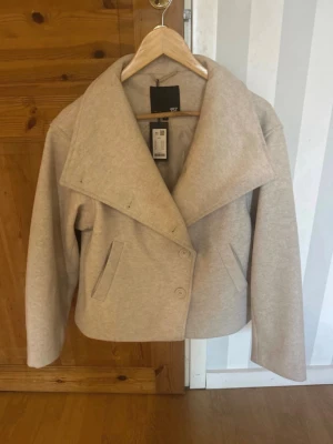 Beige kort kappa från 157 - Stilren beige kappa från 157 med bred krage och dubbelknäppning framtill. Modellen är kort och har två snedställda fickor. Perfekt för dig som gillar minimalistisk och modern stil. Materialet ser ut att vara mjukt och värmande, perfekt för kyligare dagar. Helt ny prislapp kvar