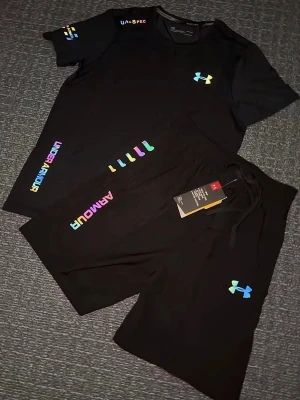 Svart träningsset från Under Armour - Svart träningsset från Under Armour med färgglada loggor och detaljer. Shorts med snörning i midjan och bekväm passform, perfekta för träning eller sportiga dagar. Materialet känns mjukt och lätt, och designen är modern med regnbågsfärgade tryck. Storlek S, helt ny 