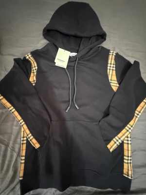 Svart Burberry hoodie med rutiga detaljer - Snygg svart hoodie från Burberry med klassiska beige-rutiga detaljer längs ärmar och axlar. Hoodien har huva med dragsko, stor magficka och logotyp på snörändarna. Perfekt för dig som vill ha en exklusiv streetwear-look med ikoniska Burberry-vibbar. Storlek M/L