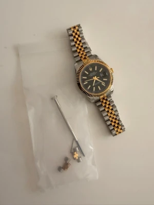  Datejust silver/guld klocka - Säljer en stilren  Datejust klocka  i tvåton (guld/silver) med svart urtavla. Klockan har en elegant design som passar både till vardag och finare tillfällen. Visare och markeringar har självlysande lume som lyser grönt i mörkret, vilket gör den lätt att läsa även på kvällen. Klockan är i 41mm 