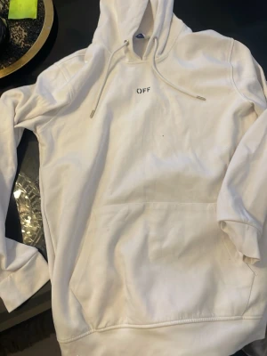 Vit hoodie från Off-White - Säljer en vit hoodie från Off-White med liten 'OFF' text på bröstet och coola svarta penseldrag på ryggen. Hoodien har huva med dragsko och stor magficka framtill. Perfekt för dig som gillar streetwear och vill sticka ut.