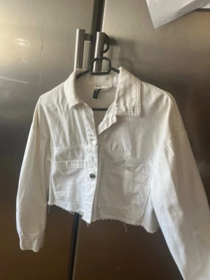 Vit croppad jeansjacka från H&M - Säljer en vit croppad jeansjacka från H&M med rå kant nertill och två stora bröstfickor. Jackan har klassisk krage, knappstängning framtill och långa ärmar. Perfekt för en trendig och avslappnad look.