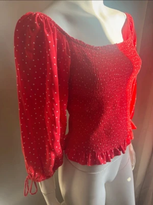 Röd prickig blus med puffärm strl M - Trendig röd blus med vita prickar, puffiga långärmar och smockad kropp. Toppen har söt volangkant nedtill och knytband vid ärmsluten. Perfekt för dig som vill sticka ut med en färgglad och feminin look.