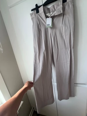 Beige vida glansiga kostymbyxor - Snygga beige kostymbyxor med hög midja och vida ben. Byxorna har pressveck framtill och stängs med dragkedja och hake. Tillverkade i ett lätt, mjukt tyg som ger en stilren glansig look. Perfekta för dig som gillar en clean och modern stil.
