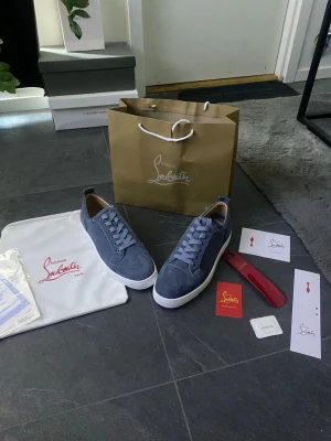 Gråa Christian Louboutin sneakers - Snygga gråa sneakers från Christian Louboutin i mocka med klassisk röd sula och vita kanter. Skorna har rund tå, matchande grå skosnören och diskret branding på insidan. Perfekta för dig som vill ha en exklusiv touch till din outfit.