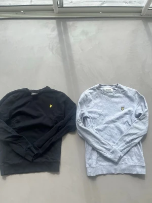 2 Lyle & Scott sweatshirts svart/grå - Två crewneck sweatshirts från Lyle & Scott, en i svart och en i ljusgrått. Båda har den klassiska gula loggan broderad på bröstet och ribbade muddar vid ärmslut och nederkant. Perfekta basplagg till garderoben.