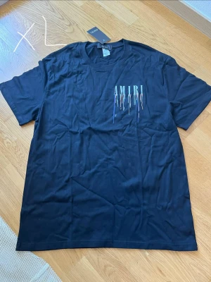 Svart AMIRI t-shirt med tryck - Säljer en svart t-shirt från AMIRI i storlek XL. T-shirten har korta ärmar och är gjord i mjuk bomull. På bröstet och ryggen finns ett stort AMIRI-tryck med färgstänk i blått och rött som ger en unik look. Perfekt för dig som gillar streetwear och vill sticka ut.