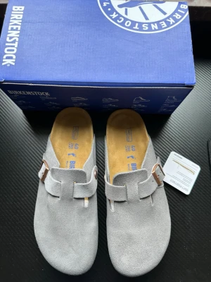 Grå mockatofflor från Birkenstock - Snygga grå tofflor från Birkenstock i mjuk mocka med klassisk slip-in design och justerbart spänne i metall. Fotbädden är i kork och innersulan är i skönt material för extra komfort. Perfekta för chill dagar hemma eller när du vill ha en clean look.
