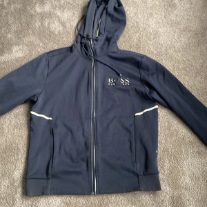 Mörkblå huvtröja från Hugo Boss - Säljer en stilren mörkblå huvtröja från Hugo Boss med dragkedja och broderad logga på bröstet. Tröjan har långa ärmar och en praktisk huva med snörning. Perfekt för en avslappnad look.