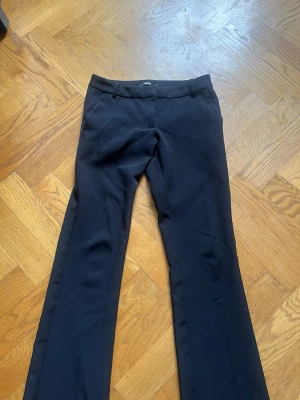 Svarta kostymbyxor bootcut - Snygga svarta kostymbyxor med bootcut passform och klassisk midja. Byxorna har hällor för skärp. Perfekta för en stilren och trendig look. Materialet känns mjukt och följsamt, troligen en blandning av polyester och viskos. 