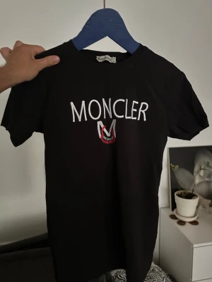 Svart Moncler t-shirt med logga - Snygg svart t-shirt från Moncler med stor vit broderad logga och emblem på bröstet. Klassisk rund halsringning och korta ärmar. Perfekt för dig som gillar stilrena och exklusiva plagg.