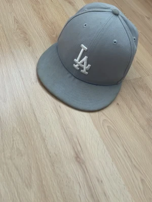 Grå LA-keps  - Snygg grå keps, LA-logga broderad framtill i vitt. Klassisk snapback-modell med rak skärm och ventilationshål. Perfekt accessoar för dig som gillar streetwear och sportig stil.