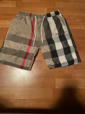 Burberry shorts - Snygga shorts med klassiskt rutigt mönster i beige, svart, vitt och rött. De har elastisk midja med vit dragsko och är tillverkade i ett lätt, glansigt material som passar perfekt till sommaren. Stilen är inspirerad av streetwear och de har en avslappnad passform.
