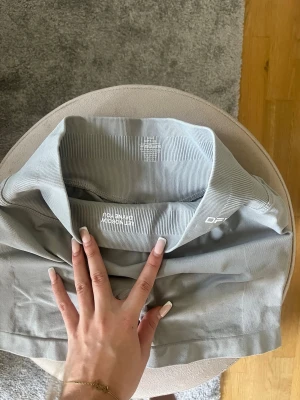 Ljusgrå shorts från Dfyne - Säljer ett par ljusgrå shorts från Dfyne med bred, ribbad hög midja. Shortsen är tillverkade i ett stretchigt syntetmaterial och har en clean, sportig look utan synliga sömmar framtill.