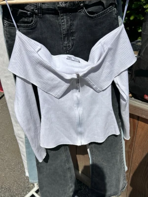 Vit ribbad offshoulder topp från Zara - Trendig vit ribbad topp från Zara med offshoulder-design och dragkedja framtill. Toppen har långa ärmar och en snygg, croppad passform som ger en cool vibe. Perfekt att styla med jeans eller kjol för en fräsch look.