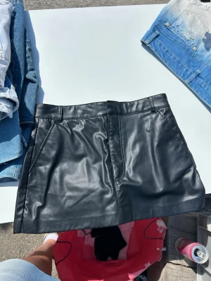 Svart skort i fuskläder - Snygg svart skort i fuskläder med fickor fram och bälteshällor i midjan. Klassisk och stilren design med shorts under kjolen för extra komfort. Perfekt för dig som vill ha en edgy look med en twist.