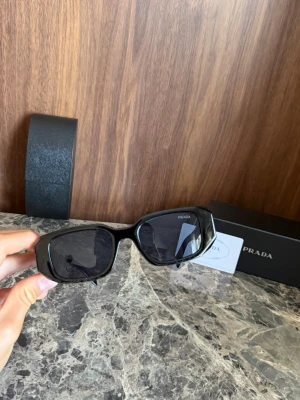Svarta solglasögon från Prada - Snygga svarta solglasögon från Prada med rektangulära bågar och mörka glas. Klassisk och stilren design med diskret Prada-logga på skalmen. Perfekta för att lyfta din look med en lyxig touch.